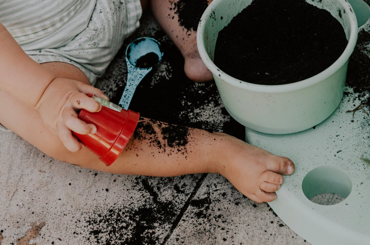 A toddler sits on the ground, playfully exploring soil with a bucket and red cup.