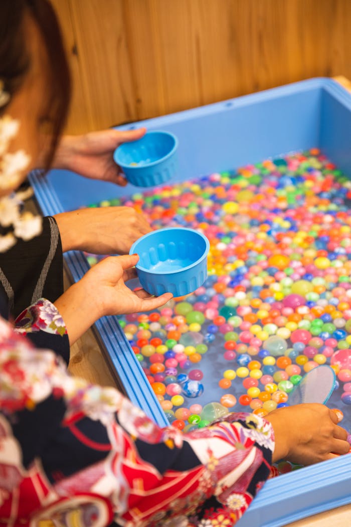 High angle anonymous females washing and rinsing multicolored bright fishbowl beads in blue basin