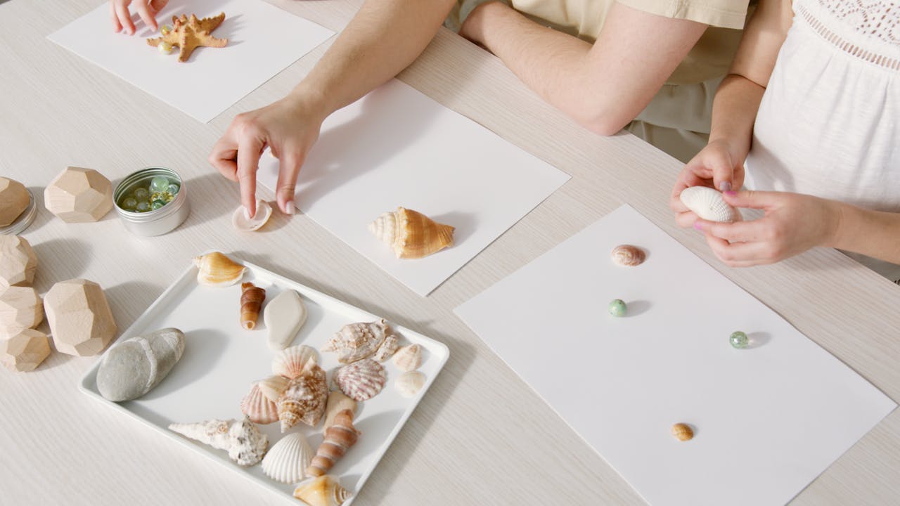 Close-up view of hands arranging seashells and wooden blocks on a white table for craft activity.