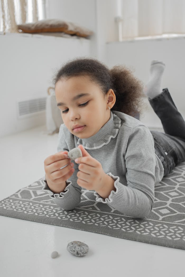 Cute little ethnic girl with dark Afro hair in casual outfit lying on floor at home and playing with natural stones during weekend